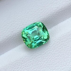 May include: A green gemstone with a cushion cut. The gemstone is set on a white background.