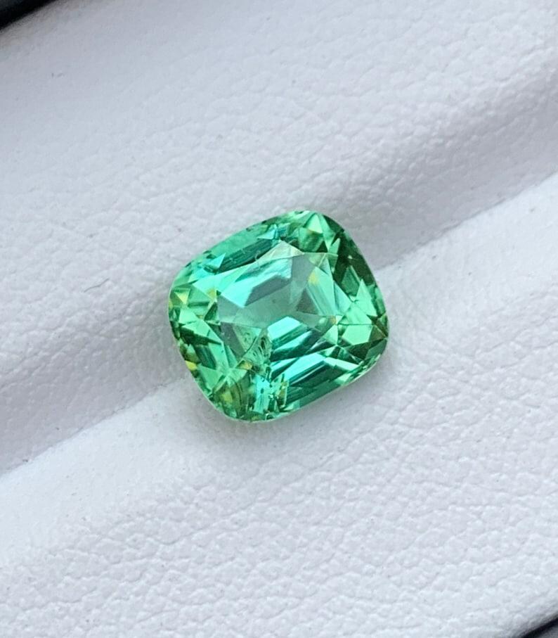May include: A green gemstone with a cushion cut. The gemstone is set on a white background.