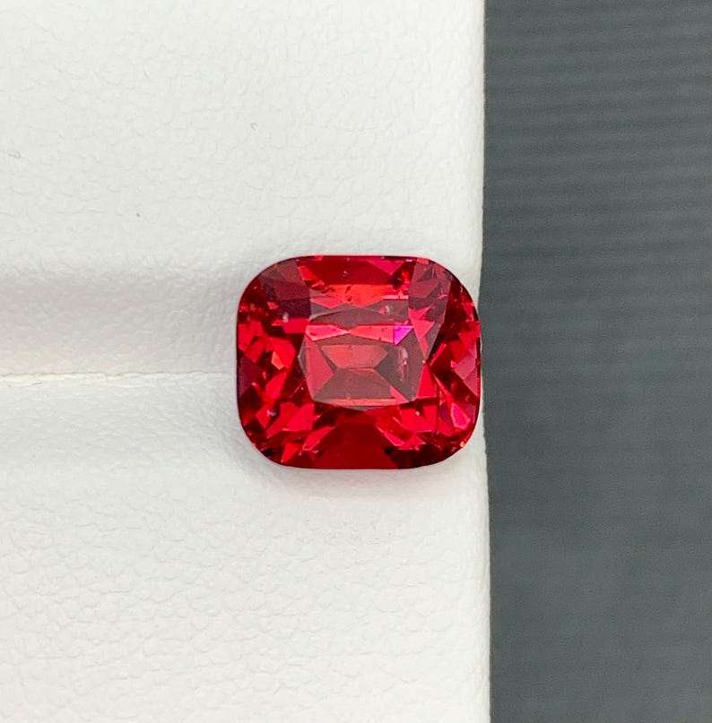 May include: A vibrant, faceted red gemstone with a cushion cut. The gem is a deep, rich red color and appears to be highly polished, reflecting light. The shape is a rounded square, showcasing the intricate internal facets.