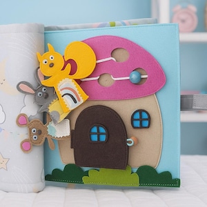 May include: A soft, interactive fabric book for babies and toddlers. The book features a light blue cover with a mushroom house design, and a gray cover with clouds, suns, and stars. Includes felt animal figures.