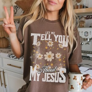「Let Me Tell You About My Jesus」シャツ、女性用クリスチャングラフィックTシャツ、デイジーがあしらわれた信仰Tシャツ、大胆な聖書の一節シャツ、礼拝シャツギフト