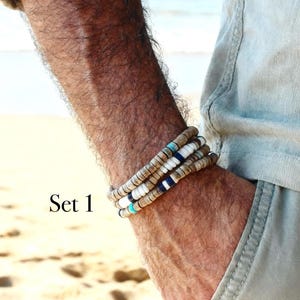 May include: Three beaded bracelets in neutral colours, with white, navy, and turquoise accents. The bracelets are stacked on a wrist, with the text "Set 1" visible. The bracelets are made of natural materials and are suitable for everyday wear.