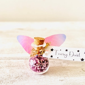 May include: A miniature glass bottle filled with pink glitter, with a cork stopper and a tag reading "Fairy Dust". The bottle is decorated with pink and purple fairy wings.