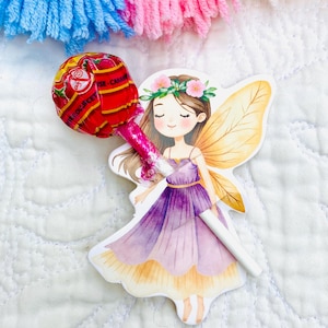 May include: A fairy-shaped sticker with a lollipop. The fairy is wearing a purple dress, a flower crown, and has golden wings. The lollipop wrapper is red with yellow details. The background is a white quilted fabric.