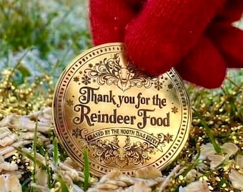 May include: A round gold-colored metal coin featuring an ornate reindeer illustration and the text "Thank you for the Reindeer Food" with "SIGNED BY THE NORTH POLE REEF" printed below, held by a hand wearing a bright red knit glove.