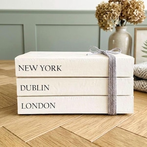 Peut inclure: Trois livres décoratifs blancs empilés, avec les noms de villes "NEW YORK", "DUBLIN" et "LONDON" imprimés. Ils sont attachés avec une ficelle grise. Un vase de fleurs séchées et une image de fougère encadrée sont en arrière-plan.