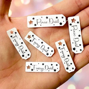 May include: Several small, rectangular wooden tags with a white wood grain effect. The tags display the words "Fairy Dust" or "Pixie Dust" in black script, with star-shaped cutouts and small black star details. Ideal for craft projects or adding a magical touch to gifts.