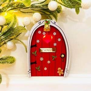 May include: A festive red fairy door with a white frame, decorated with snowflakes, holly, and a gingerbread man. It has a gold bell and the text "ELF WORKSHOP". A glowing red heart is in the center.