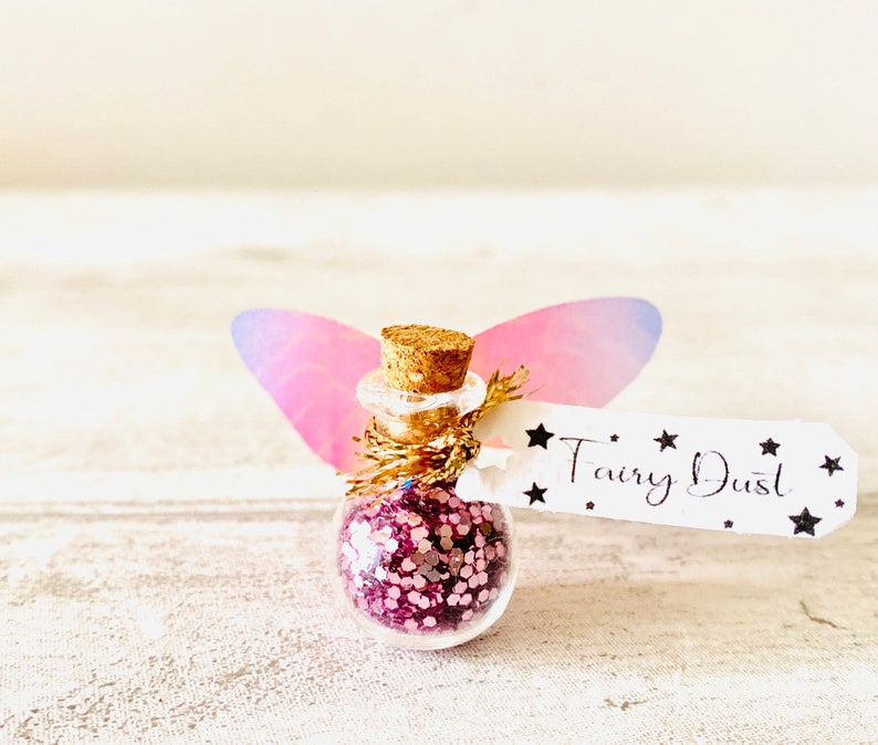 May include: A miniature glass bottle filled with pink glitter, with a cork stopper and a tag reading "Fairy Dust". The bottle is decorated with pink and purple fairy wings.