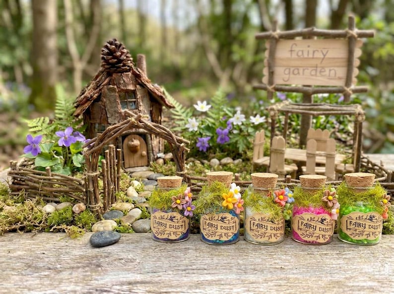 Pode incluir: Uma cena de jardim de fadas em miniatura com uma casa de madeira r&uacute;stica, uma placa que diz "fairy garden" e cinco pequenos frascos de vidro rotulados como "fairy dust". Os frascos s&atilde;o decorados com musgo e flores.