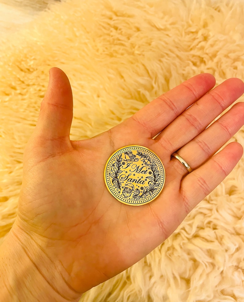 Puede incluir: Una moneda navide&ntilde;a dorada con el texto "I Met Santa" en una fuente decorativa. La moneda presenta un dise&ntilde;o de corona con una ilustraci&oacute;n de trineo y Pap&aacute; Noel. La moneda se sostiene en una mano.