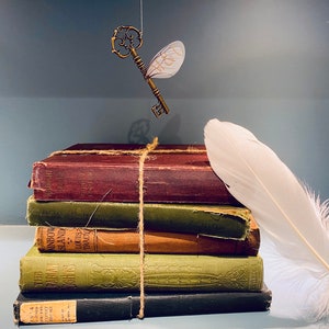 May include: A stack of five vintage books tied together with twine, a white feather, and a golden key with wings hanging above. The books have brown, green, and black covers with text on the spines.