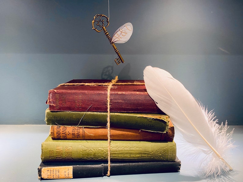 May include: A stack of five vintage books tied together with twine, a white feather, and a golden key with wings hanging above. The books have brown, green, and black covers with text on the spines.