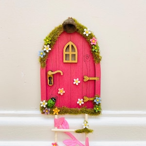 May include: A bright pink fairy door with a wooden arched window and a small ladder. The door is decorated with faux moss, colourful flowers, and a ladybug. The door has a golden handle and a keyhole.