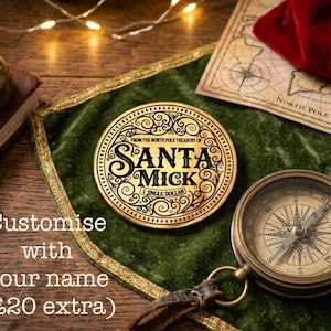 Puede incluir: Una moneda dorada con el texto "SANTA MICK" y "1 JINGLE DOLLAR" sobre una superficie de terciopelo verde, junto a una br&uacute;jula y un mapa. La moneda forma parte del "North Pole Treasury". La imagen tambi&eacute;n incluye el texto "Customise with your name".