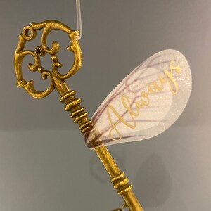 May include: A gold-colored key with intricate details and a delicate, white, translucent wing with the word "Always" written in gold lettering.