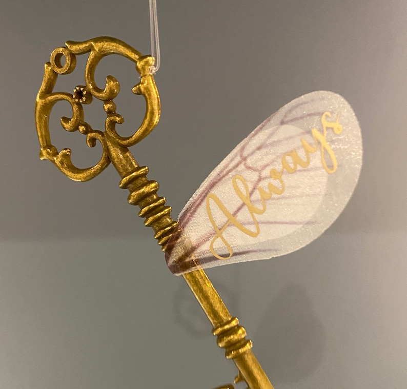 May include: A gold-colored key with intricate details and a delicate, white, translucent wing with the word "Always" written in gold lettering.