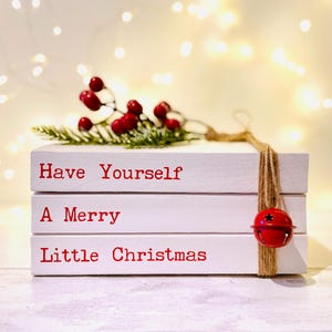 Puede incluir: Bloques de madera blancos apilados con el texto "Have Yourself A Merry Little Christmas" en rojo. Una ramita de bayas y una campana roja con una cuerda de yute añaden detalles festivos. Decoración navideña.