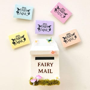 May include: A white wooden fairy mail post box with the words "FAIRY MAIL" in brown. The box is decorated with moss, flowers, and miniature mushrooms. Above the box are five colorful rectangles with the words "SPECIAL DELIVERY FAIRY MAIL".