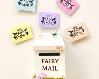 May include: A white wooden fairy mail post box with the words "FAIRY MAIL" in brown. The box is decorated with moss, flowers, and miniature mushrooms. Surrounding the box are five colorful rectangular cards with the text "SPECIAL DELIVERY FAIRY MAIL".