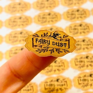 Op de afbeelding: Een vel met ronde, goudkleurige stickers met de tekst "Fairy Dust" in een zwart, gestileerd lettertype. De stickers bevatten een zwart silhouet van een heks die op een bezem vliegt met een zwarte kat.
