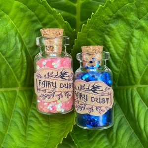 May include: Two small glass bottles with cork stoppers filled with pink and blue glitter. Both bottles have a brown paper label that reads "Fairy Dust".