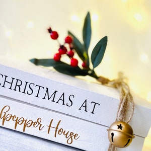 May include: Two white decorative books stacked with the words "CHRISTMAS AT Culpepper House" printed on them. A sprig of red berries and green leaves rests on top, with a gold bell hanging from twine.