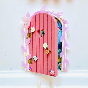 以下が含まれることがあります： ハート型の切り抜きがあるピンク色の妖精のドア。花と葉で飾られています。ドアは開いており、カラフルなシーンが見えます。小さな木製の梯子が下に置かれています。装飾的なポンポンが上に吊り下げられています。