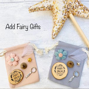 Peut inclure: Un ensemble cadeau sur le th&egrave;me des f&eacute;es avec une baguette en forme d'&eacute;toile et deux sacs &agrave; cordon. Chaque sac contient un disque en bois avec du texte, une fleur, une loupe, une aile de f&eacute;e et d'autres petits bibelots. Le texte "Add Fairy Gifts" est visible.