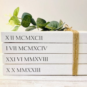 Peut inclure: Une pile de quatre livres blancs avec des chiffres romains et des années imprimés sur la face avant. Les livres sont liés ensemble avec de la ficelle et une brindille de verdure est placée sur le dessus.