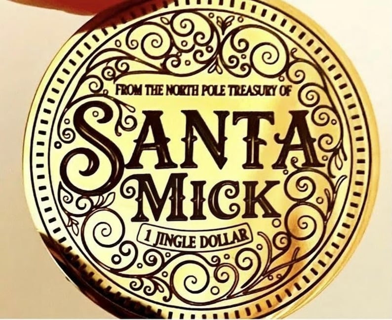 Puede incluir: Una moneda dorada con las palabras "SANTA MICK" en una fuente decorativa. La moneda tambi&eacute;n presenta el texto "FROM THE NORTH POLE TREASURY OF" y "1 JINGLE DOLLAR". La moneda tiene un borde adornado con volutas.