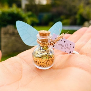 May include: A small glass bottle filled with gold glitter, labeled "Fairy Dust." The bottle has a cork stopper, gold twine, and light blue paper wings attached. The background is blurred green foliage.