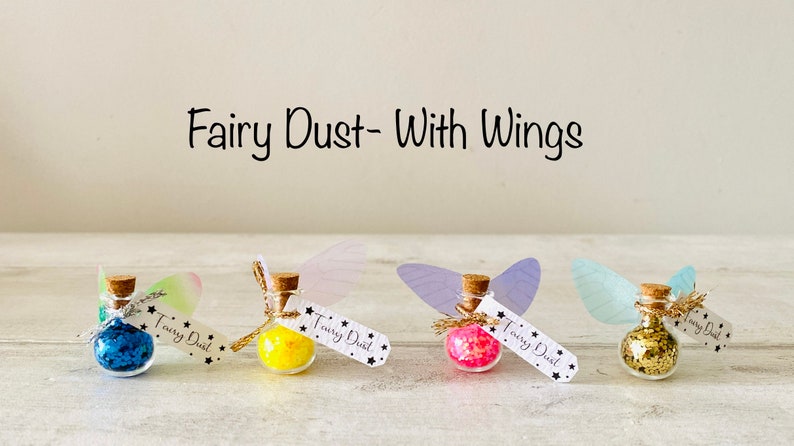 May include: Four small glass bottles filled with colorful glitter, each with a cork stopper and a tag that reads "Fairy Dust". Each bottle has a pair of attached wings. The text "Fairy Dust - With Wings" is above the bottles.