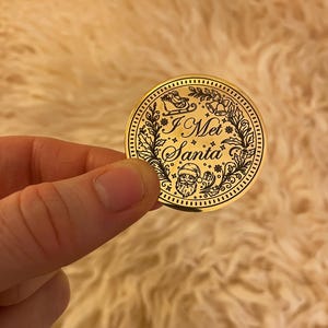 Puede incluir: Una moneda dorada de Navidad con la inscripci&oacute;n "I Met Santa". La moneda presenta un dise&ntilde;o detallado de Pap&aacute; Noel, un trineo, campanas y follaje festivo. La moneda est&aacute; sostenida en una mano.