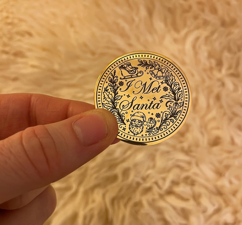 Puede incluir: Una moneda dorada de Navidad con la inscripci&oacute;n "I Met Santa". La moneda presenta un dise&ntilde;o detallado de Pap&aacute; Noel, un trineo, campanas y follaje festivo. La moneda est&aacute; sostenida en una mano.