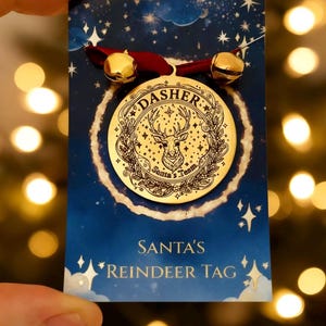 Puede incluir: Adorno navideño dorado con la palabra "DASHER" y un diseño de reno. El adorno está sujeto a una tarjeta azul con el texto "SANTA'S REINDEER TAG" y dos campanas doradas.