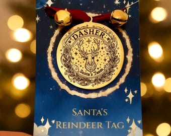 May include: A gold-colored circular metal tag featuring an engraved reindeer head, the text "DASHER" and "Santa's Team," attached to a red ribbon with two small gold jingle bells, mounted on a blue card labeled "SANTA'S REINDEER TAG."
