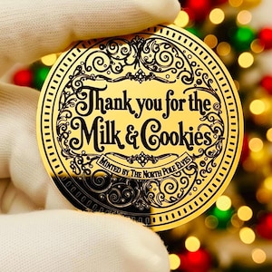 以下が含まれることがあります： 「Thank you for the Milk & Cookies」の黒い文字が書かれた、金色の丸いコイン。手袋をした手で持っており、背景にはぼやけたクリスマスツリーがあります。コインは「Minted by the North Pole Elves」です。
