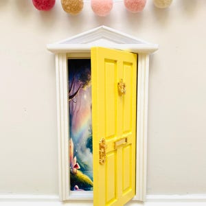 May include: A small, bright yellow door with gold fittings is ajar, showing a vibrant fantasy landscape. The door is set in a white frame, with a garland of pastel pom-poms above. A whimsical decorative item.
