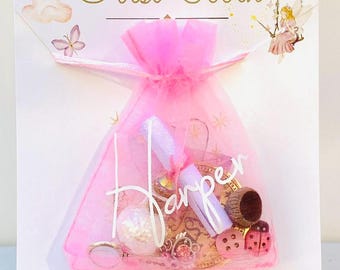 May include: A pink organza gift bag filled with small toys and trinkets, with the name "Harper" in white script. The bag is tied with a pink ribbon and sits on a card with the words "First Tooth" in gold script, along with fairy illustrations.