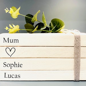 May include: A stack of four white wooden blocks with the words "Mum", a heart, "Sophie", and "Lucas" written on them. The blocks are tied together with a piece of twine and decorated with a sprig of green leaves.