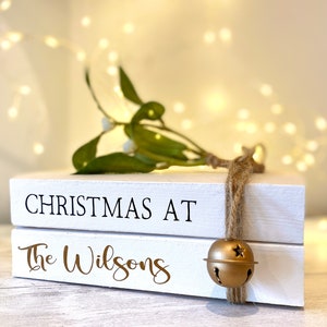 Puede incluir: Dos bloques de madera blancos apilados con las palabras "CHRISTMAS AT" y "The Wilsons". Una campana dorada cuelga de una cuerda de hilo. Muérdago verde con bayas blancas encima. Luces de hadas en el fondo.