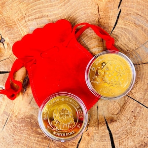 Peut inclure: Deux pièces de vœux dorées du Père Noël dans des étuis de protection transparents, à côté d'une pochette en velours rouge. Une pièce représente le Père Noël, l'autre le traîneau. Les pièces sont sur une surface en bois.