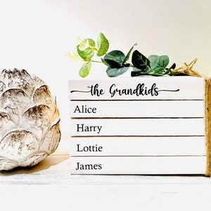 May include: A stack of white books with the text "The Grandkids" on the top book. The names Alice, Harry, Lottie, and James are written on the books below. The books are tied together with twine and a sprig of greenery is placed on top.