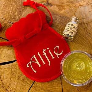Könnte beinhalten: Ein roter Samtbeutel mit dem Namen "Alfie" in weißer Schrift, eine kleine Glasflasche mit der Aufschrift "Elf Dust" und eine goldfarbene Münze mit dem Text "Santa Claus Wishing Coin". Die Gegenstände befinden sich auf einer Holzoberfläche.