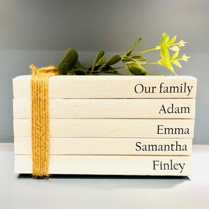 May include: A stack of five white wooden blocks with the words "Our family", "Adam", "Emma", "Samantha", and "Finley" written on them. The blocks are tied together with twine and decorated with a sprig of green leaves.