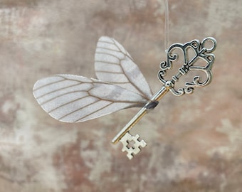 May include: A silver key with intricate details and delicate, white wings attached. The key is suspended from a string against a light brown background.