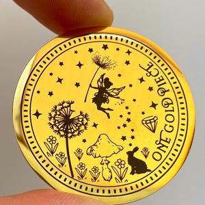 Pode incluir: Uma moeda dourada com um design gravado detalhado. A moeda apresenta uma fada, um dente-de-le&atilde;o, um cogumelo, um coelho e estrelas. O texto "ONE GOLD PIECE" est&aacute; inscrito ao redor da borda. A moeda &eacute; segurada pelos dedos de uma pessoa.