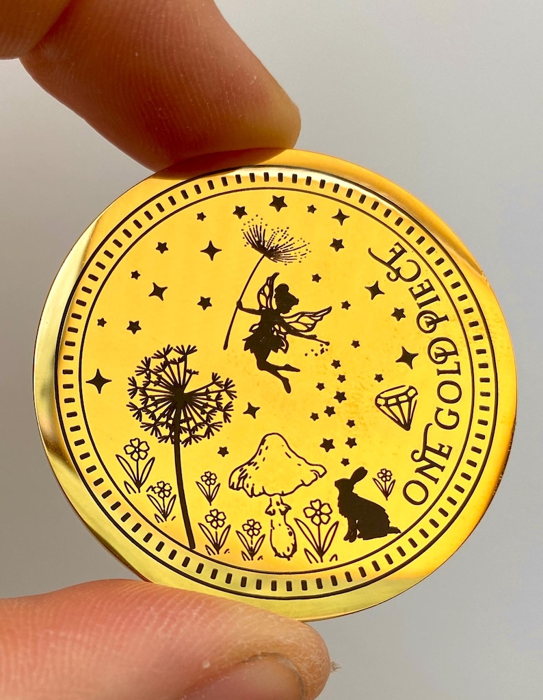 Pode incluir: Uma moeda dourada com um design gravado detalhado. A moeda apresenta uma fada, um dente-de-le&atilde;o, um cogumelo, um coelho e estrelas. O texto "ONE GOLD PIECE" est&aacute; inscrito ao redor da borda. A moeda &eacute; segurada pelos dedos de uma pessoa.