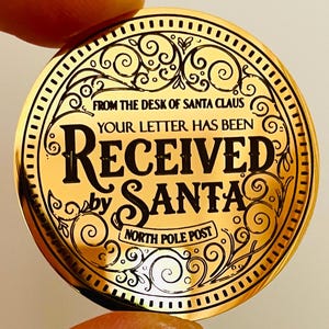 Puede incluir: Una moneda circular de metal dorado con texto grabado en negro que dice FROM THE DESK OF SANTA CLAUS, YOUR LETTER HAS BEEN RECEIVED by SANTA y NORTH POLE POST, rodeada de intrincados patrones de filigrana en color negro.